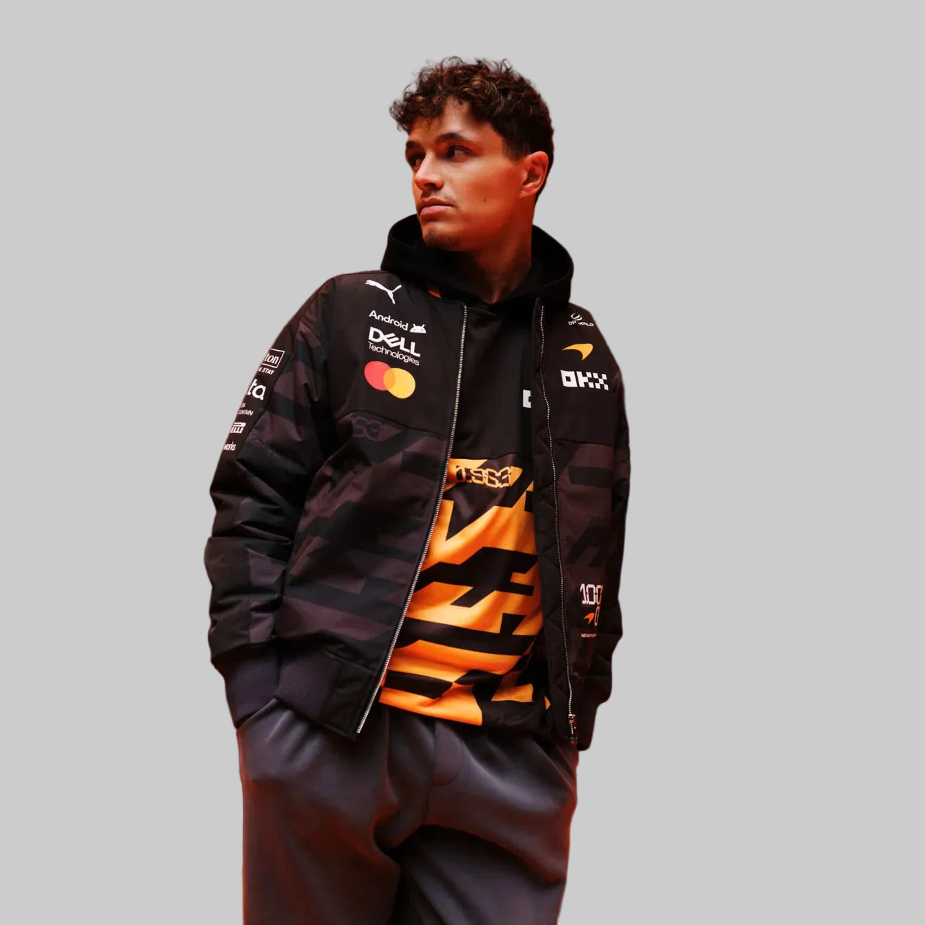 Front view of the black and gray McLaren inspired 1000th GP unisex bomber jacket with multi-sponsor logos, orange striped collar, and 1000 GP badge by TassFit Performance.